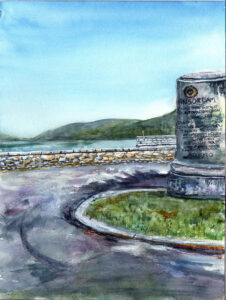 Curved road with grassy median and stone monument by a low wall near water and hills.